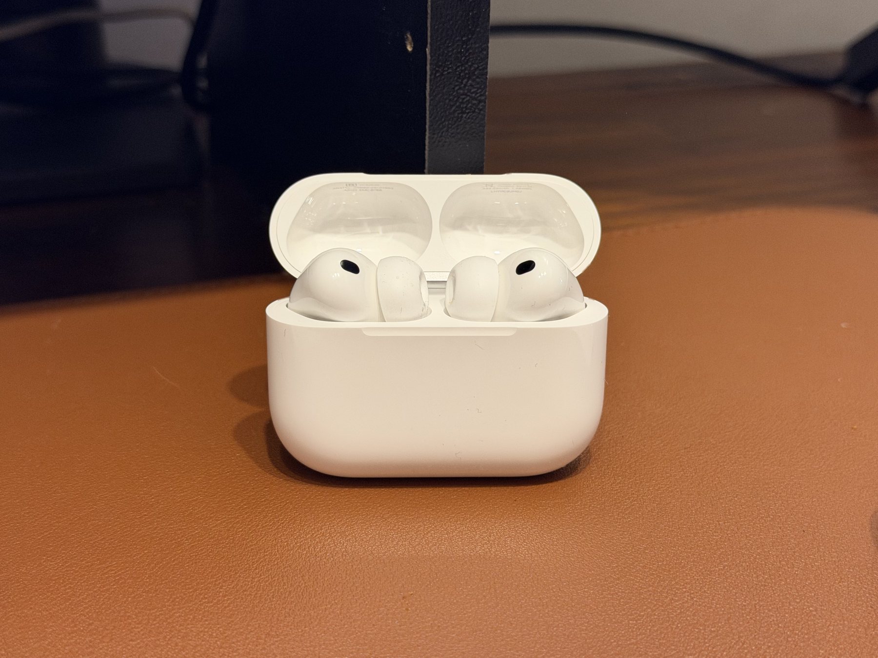 White AirPods Pro 3 sitting in their open charging case on a brown leather desk mat